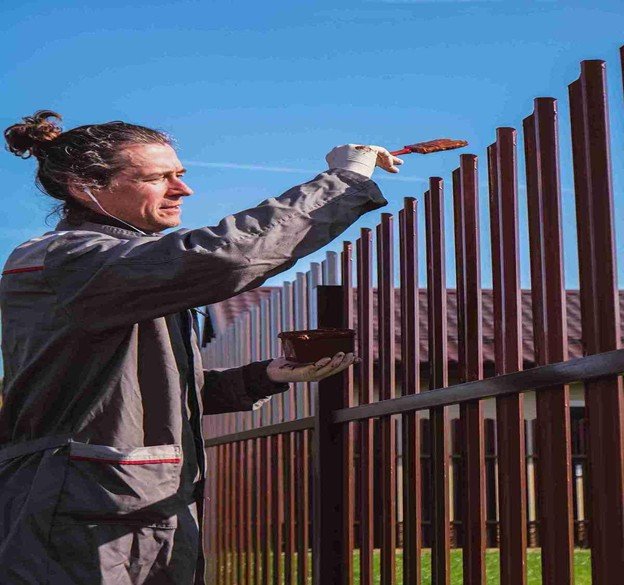 A worker Cedar fence board fastening with a brush while holding a container of paint outdoors.